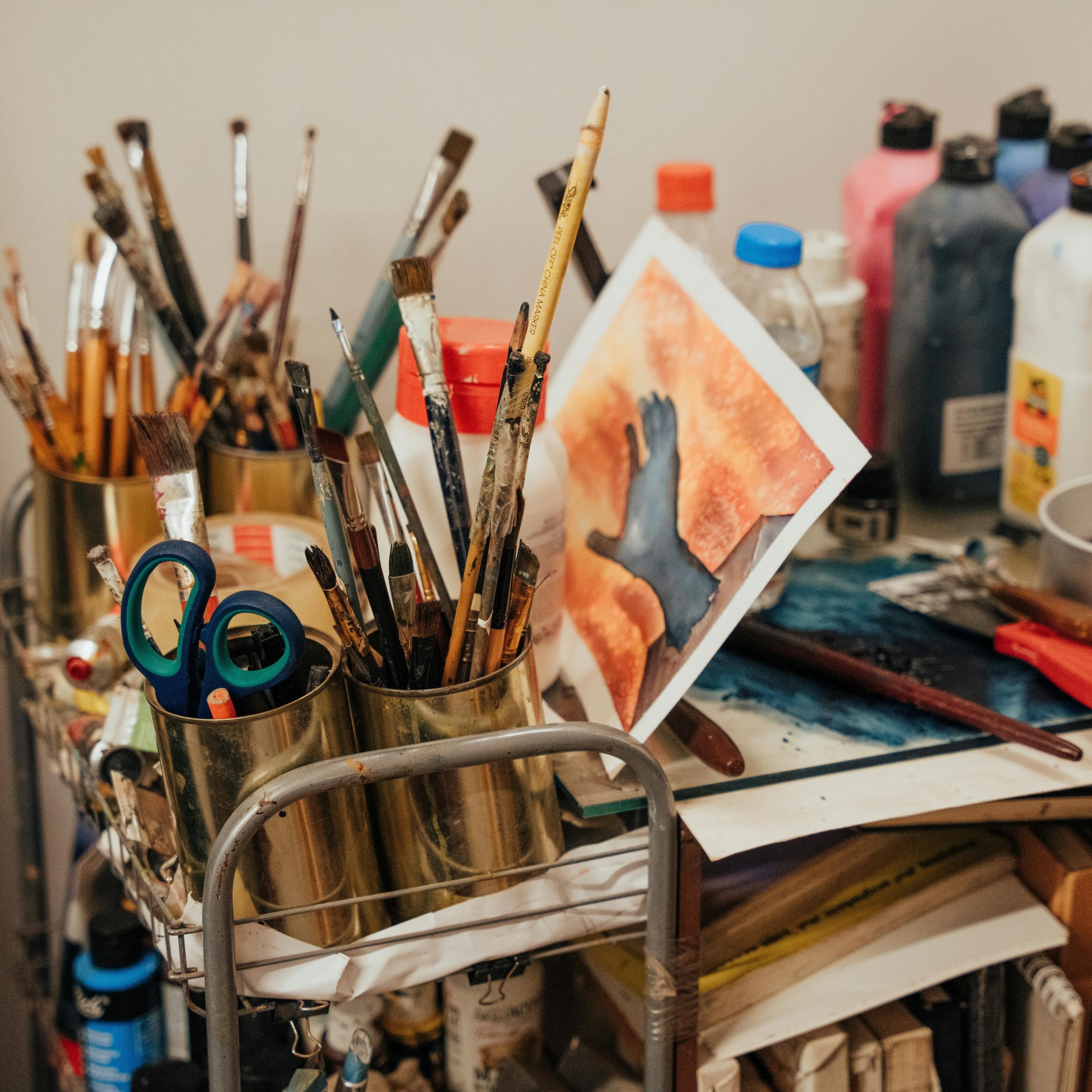 Art supplies including paintbrushes in containers, scissors, paint bottles, and artwork on cluttered desk surface.