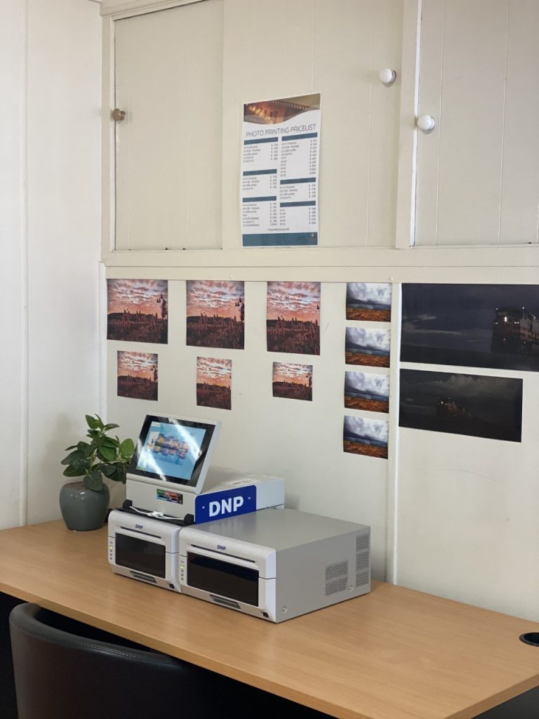 White cabinets, wall-mounted photos, wooden desk, white printer, small plant, blue DNP box.